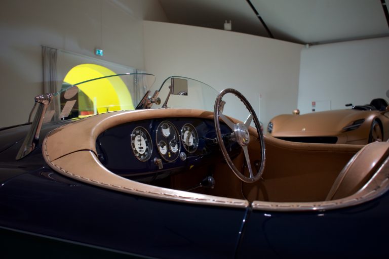 Interior view of a vintage car featuring a wooden steering wheel, classic gauges, and tan leather accents, with another sleek, modern car visible in the background.