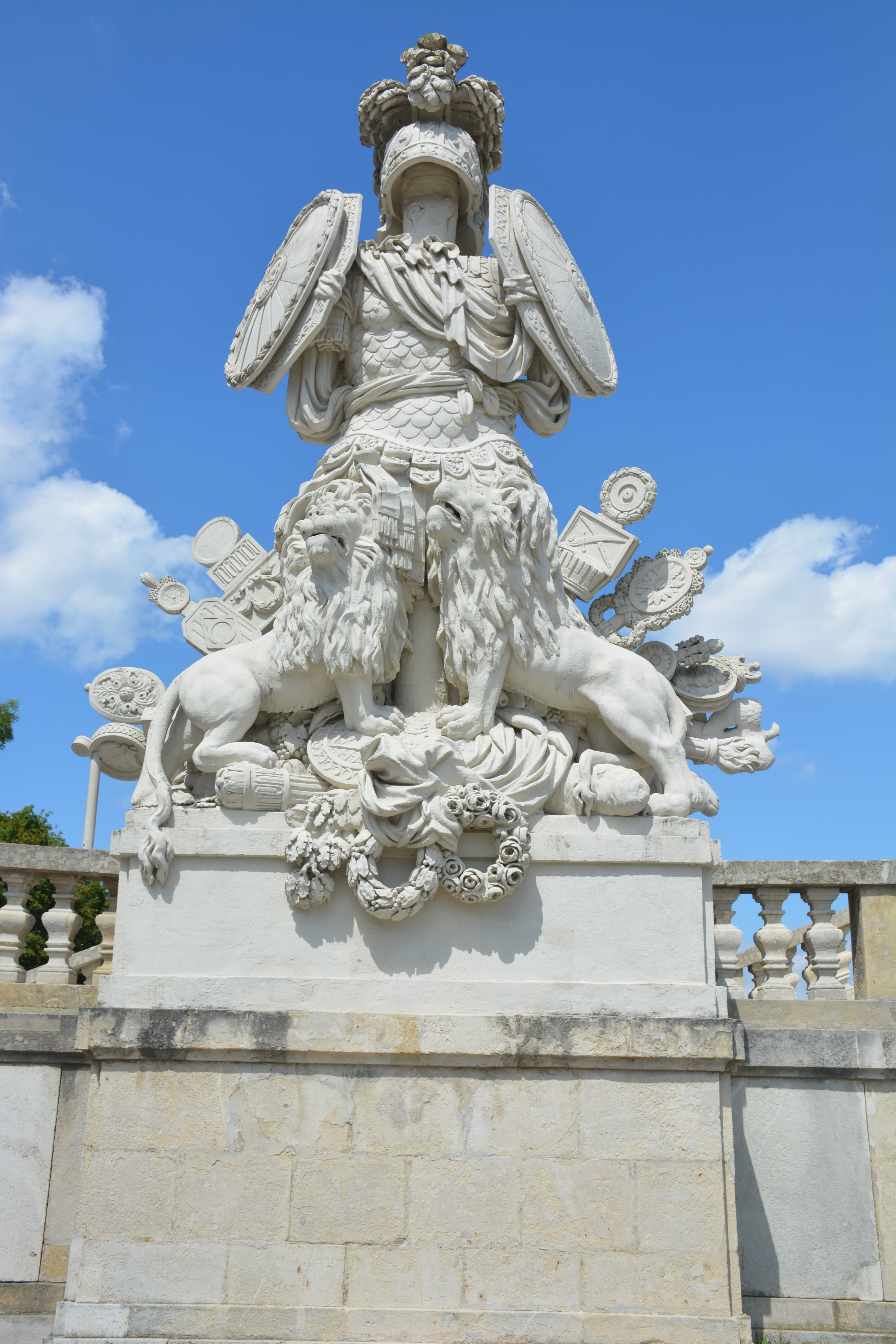 A white stone sculpture featuring a figure in armor holding shields, flanked by two lions, and topped with a crown, set against a blue sky with some clouds. The sculpture is positioned on a stone pedestal with decorative elements. It is a guardian statue at the Schönbrunn Gloriette, Vienna, Austria.