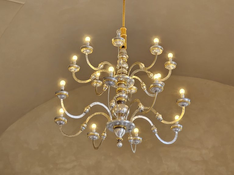 A multi-tiered chandelier with silver and gold accents against a light beige or cream-colored ceiling part of Prague Castle. The chandelier has multiple curved arms extending from a central stem, each holding a light bulb.