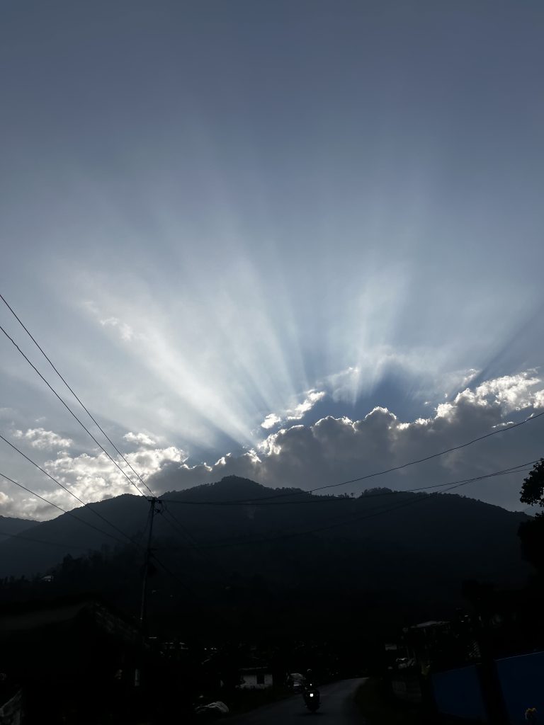 A silhouette of a mountain range with rays of sunlight streaming through the clouds above, illuminating the sky.