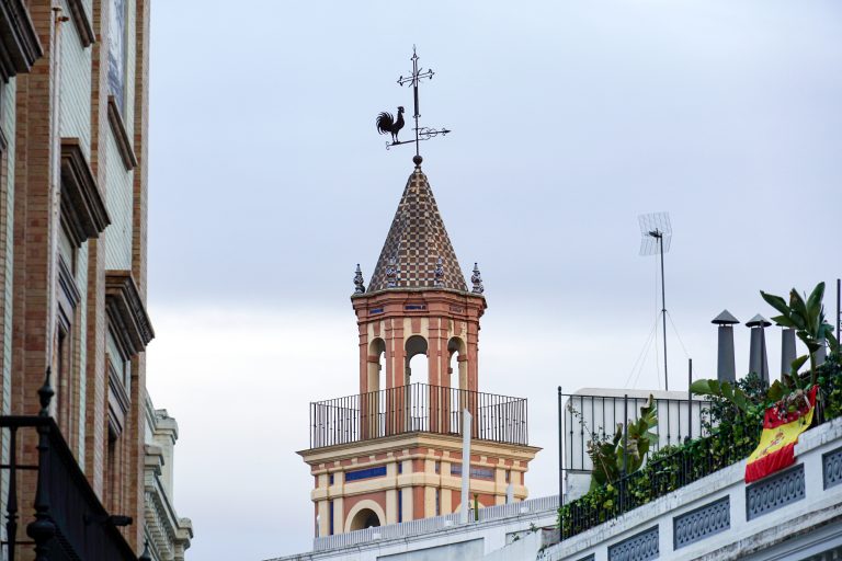 A tall, ornate tower with a checkered conical roof and a rooster-shaped weather vane on top, situated among buildings. The foreground includes a balcony with plants and a Spanish flag draped over the railing.