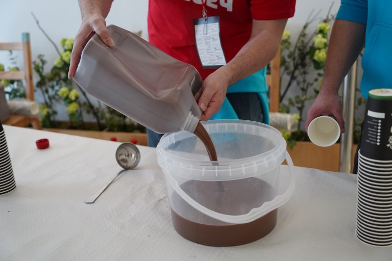 A person pouring hot chocolate from a large plastic jug into a clear plastic container on a table. Another person holds a paper cup nearby. Disposable cups and a ladle are visible on the table.