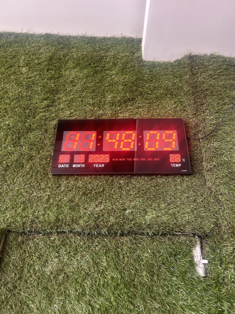 A digital clock with red LED display sits on a patch of artificial grass. It shows numerical placeholders for time, date, month, and year, alongside indicators for the days of the week and temperature.