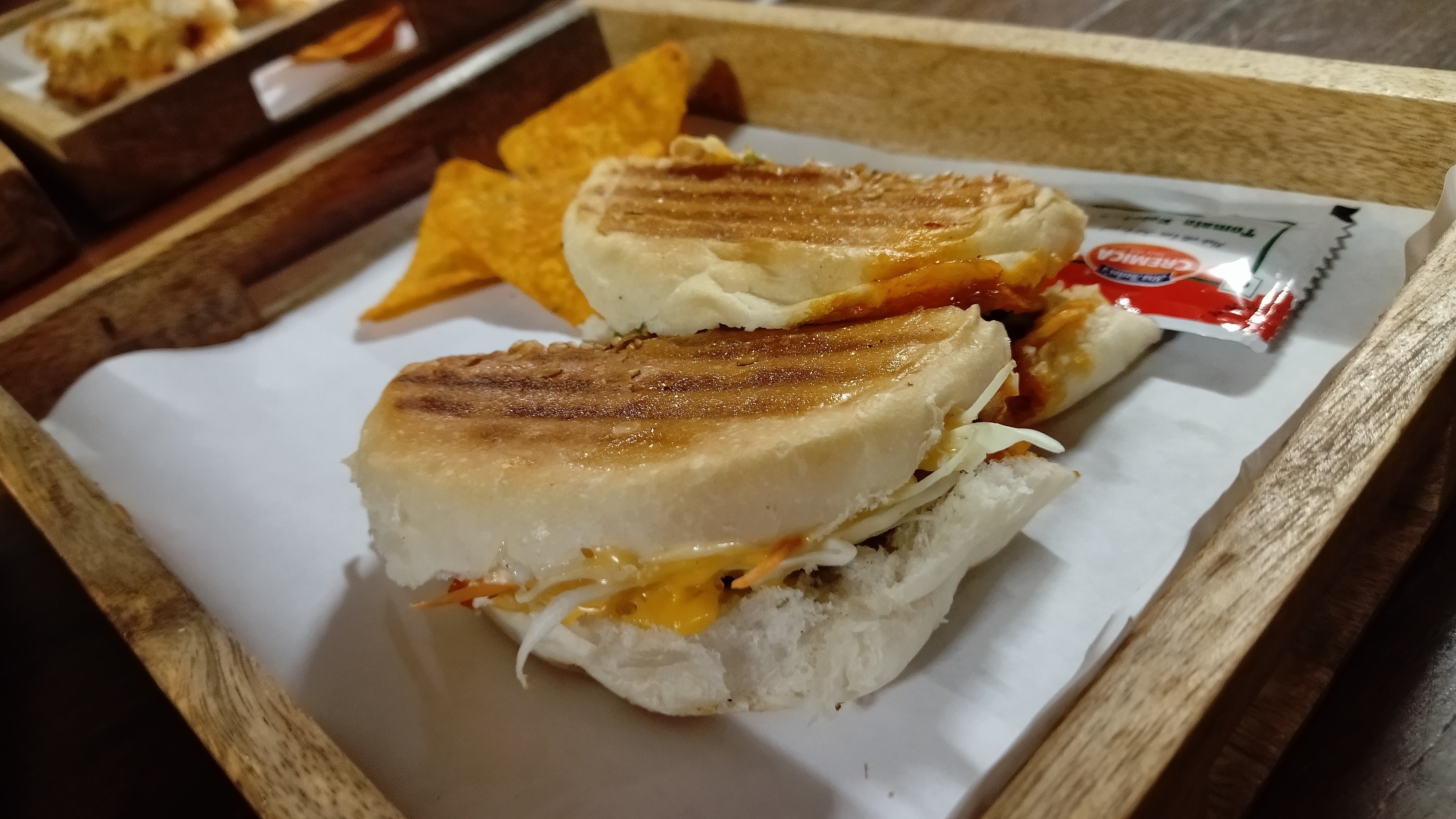 A grilled sandwich filled with shredded cabbage, cheese, and sauce, served in a wooden tray with triangular corn chips and a packet of ketchup.