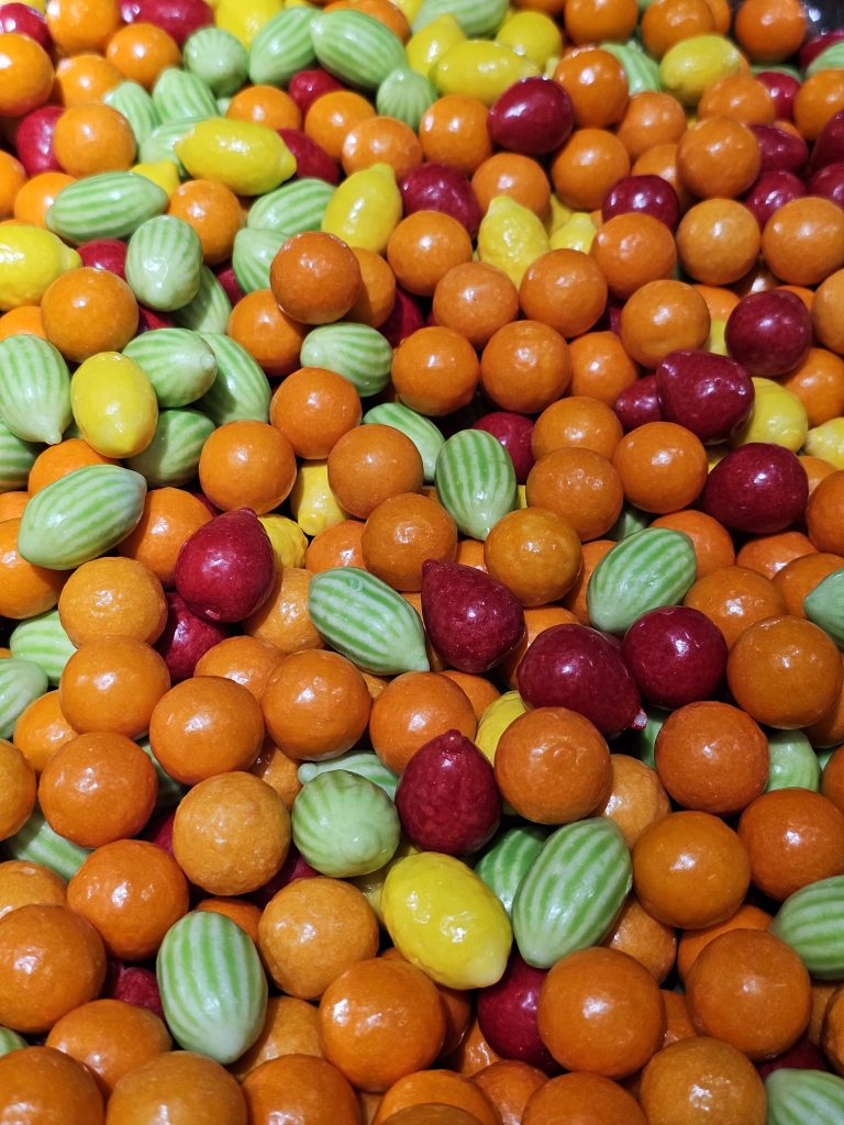 A close-up view of a mix of colorful candies, including orange, red, yellow, and green. The candies are in the shape of vegetables or fruits.