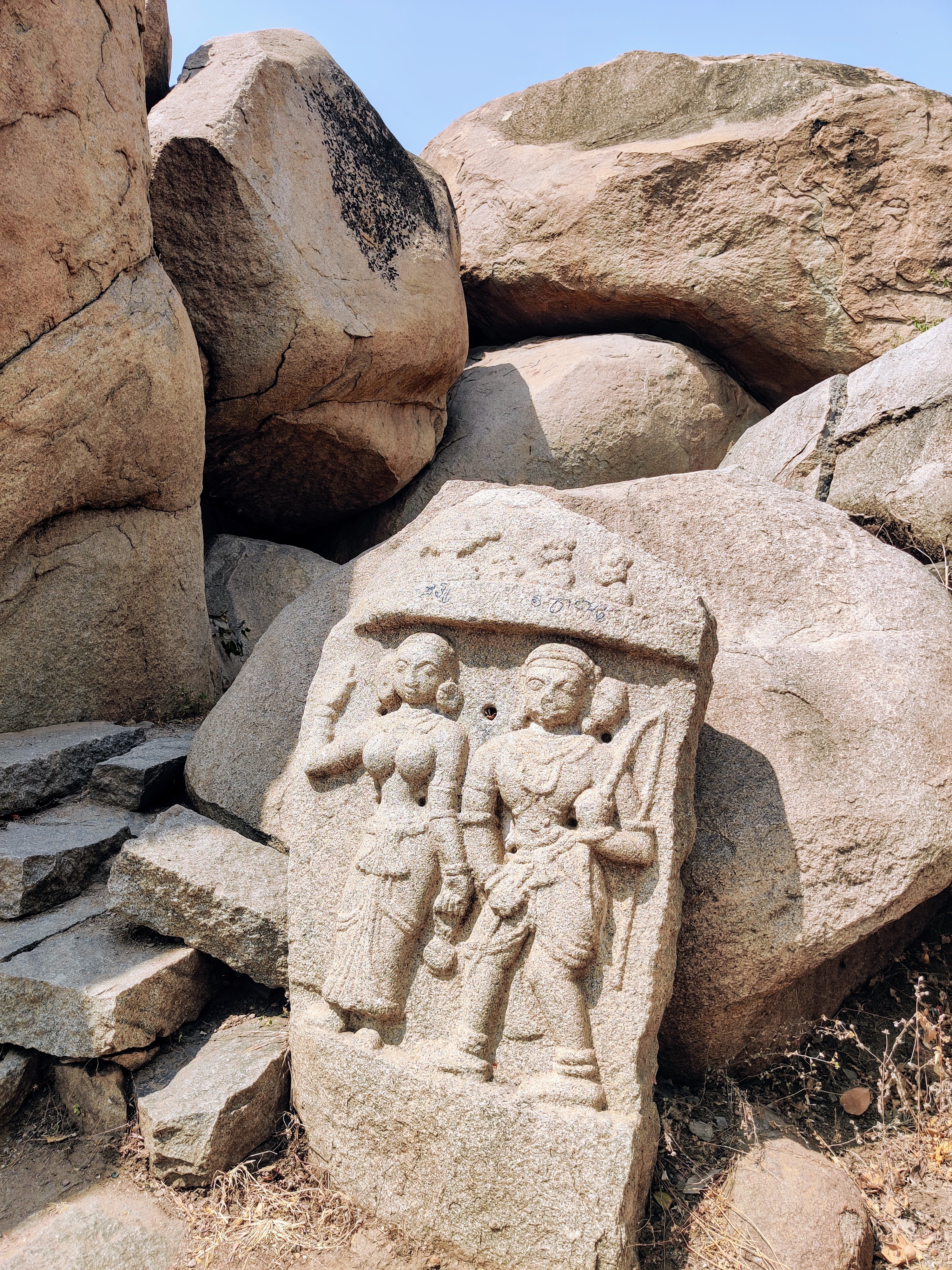 Ancient stone carving depicting two figures standing side by side, one holding a weapon and the other raising a hand, set against a backdrop of large rocks (Lord Ram, Seetha).