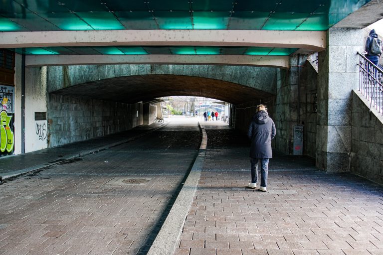 A person wearing a dark coat walks through an underpass with a textured stone ceiling and walls. The path is lined with graffiti-covered walls and is lighted by green-tinted lights.