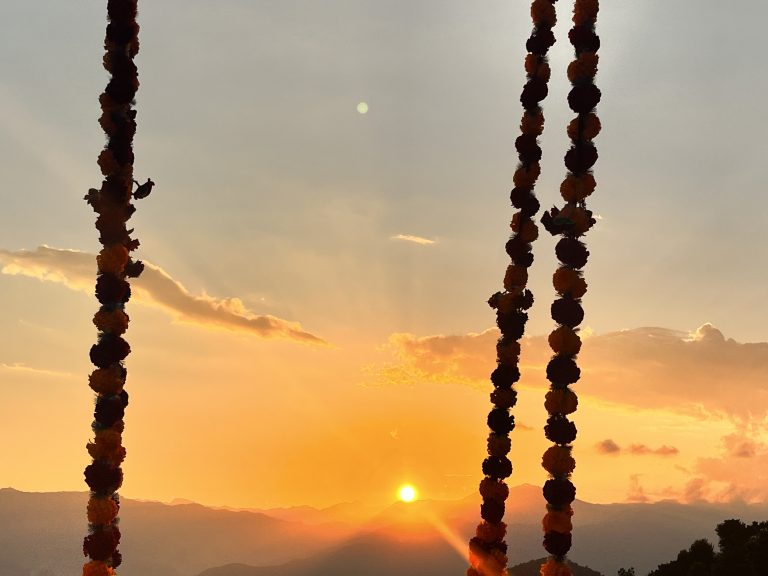 A sunset scene with the sun low on the horizon casting an orange and pink glow across the sky. In the foreground, three vertical garlands of flowers create silhouettes against the vibrant background. The landscape beneath includes silhouetted hills and soft clouds dispersed across the sky.