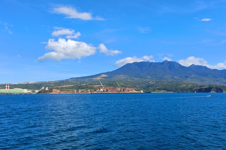 A scenic view of a deep blue ocean with a coastline featuring a construction site and large industrial structures. In the background, a lush green mountain range under a bright blue sky with scattered white clouds. Camaya Coast, Philippines