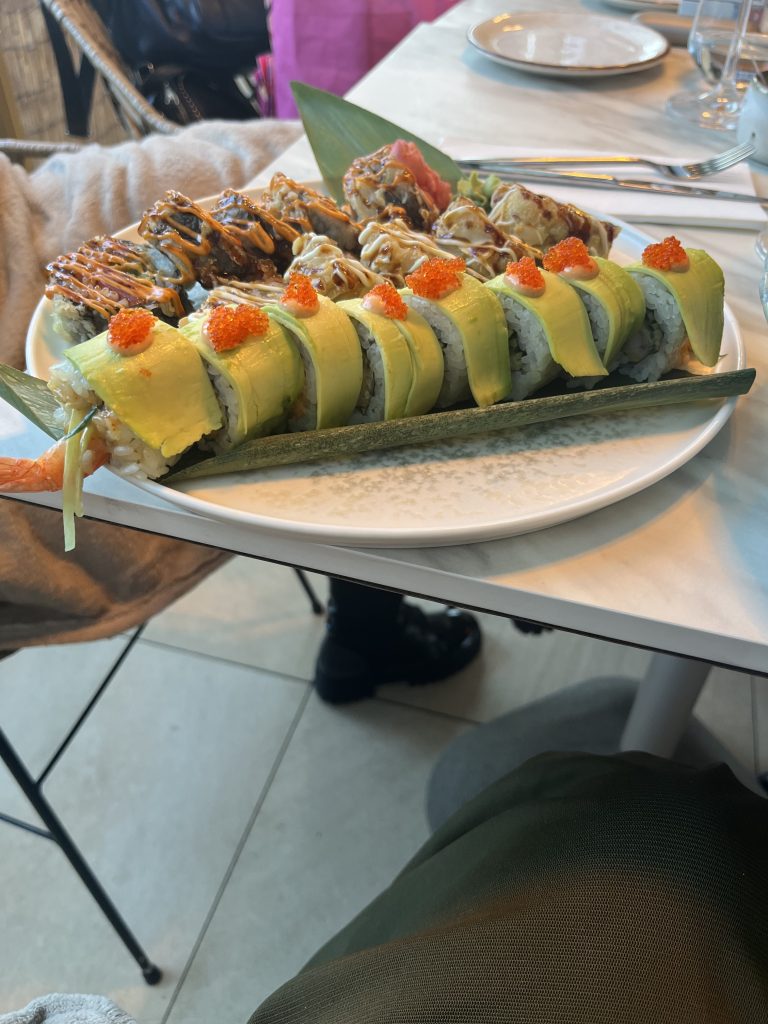 A plate of assorted sushi rolls on a white table. The rolls include slices of avocado and are topped with orange fish roe and drizzled with sauces. There are decorative leaf garnishes on the plate.