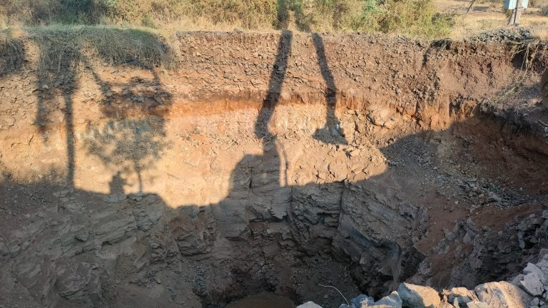 An excavation site with rugged rock formations and exposed soil layers, illuminated by warm sunlight. The shadows of construction equipment loom over the pit, highlighting the depth and scale of the dig.