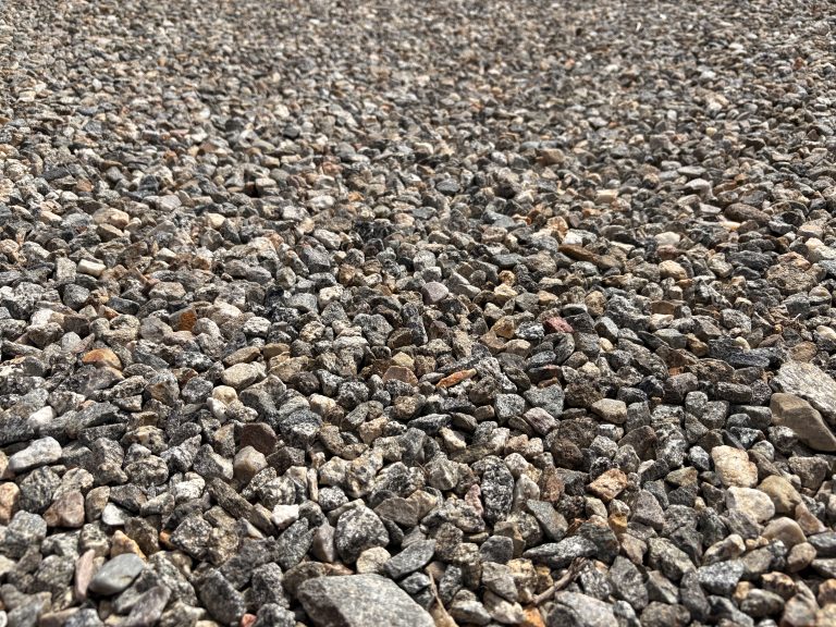 This closeup of crushed gravel shows a dense surface of small, irregularly shaped gray and tan stones. Various sized rock fragments create a textured pattern extending across the entire frame. The predominantly gray aggregate includes scattered beige and reddish-brown pieces, giving subtle color variation to the otherwise monochromatic surface.