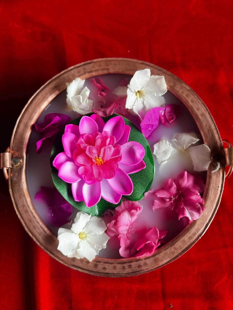 A decorative bowl with a mix of flowers floating in water, including a prominent pink lotus in the center, surrounded by white and pink petals, on a red fabric background.