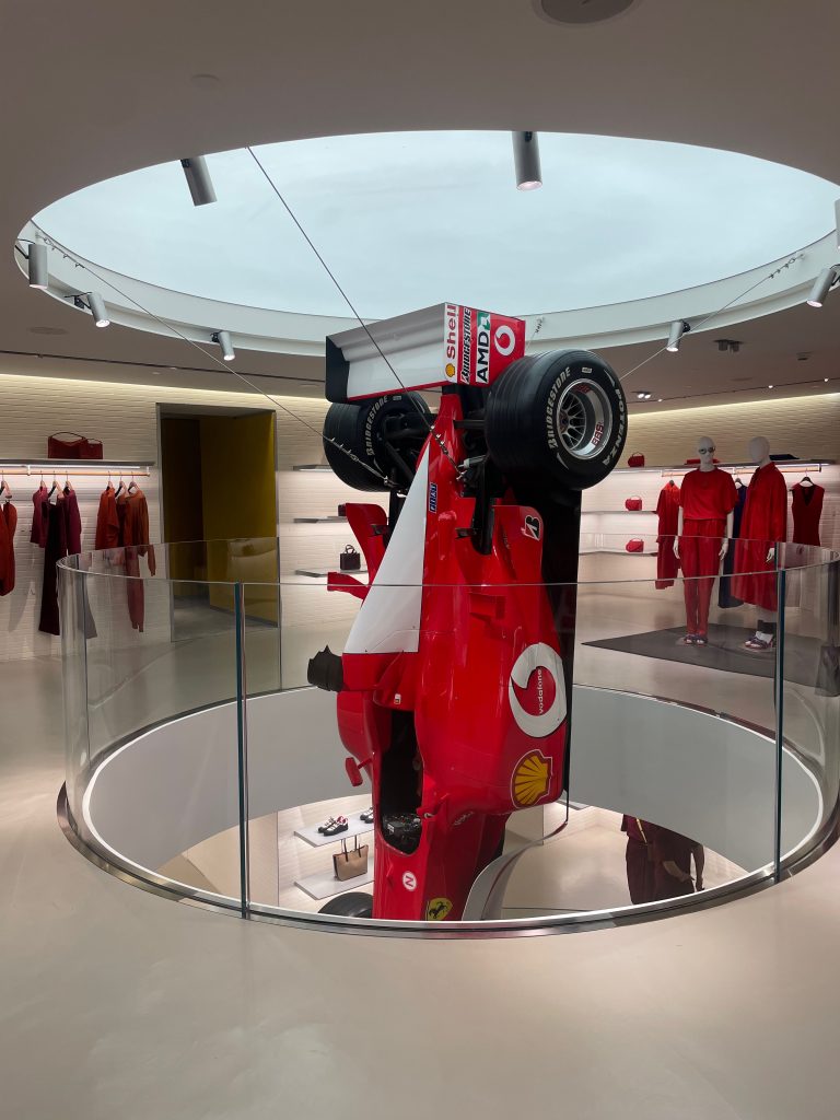 A red Formula 1 car, driven by Michael Schumacher and suspended vertically in a modern store display, with racing suits and merchandise in the background.