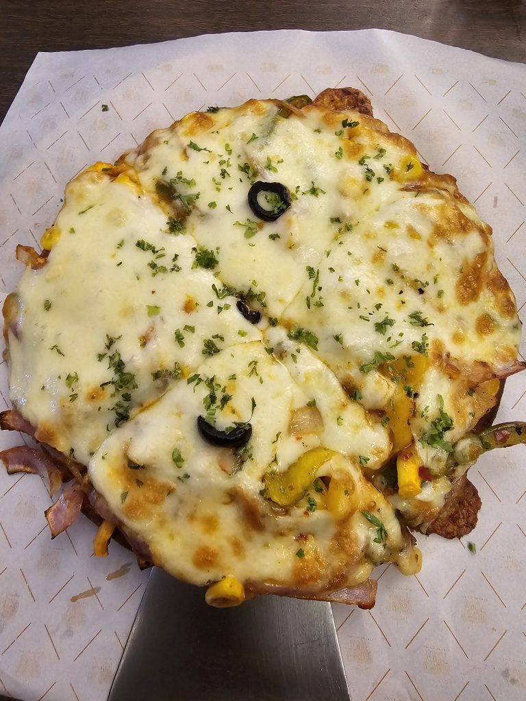 A pizza with melted cheese, toppings like olives and herbs, and a crispy crust. The pizza is served on a spatula and placed on a patterned paper.