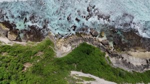 Aerial view from a cliff south of Uluwatu Temple in Bali, showing rugged coastal terrain where green vegetation meets a rocky shoreline.