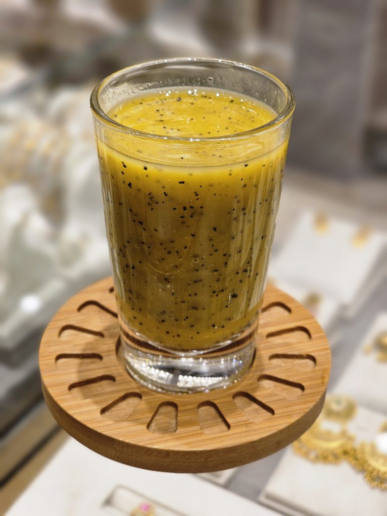 A glass of passion fruit smoothie (yellow-orange, with black seeds) on a wooden coaster against a blurred store background. Captured in Kozhikode, Kerala.