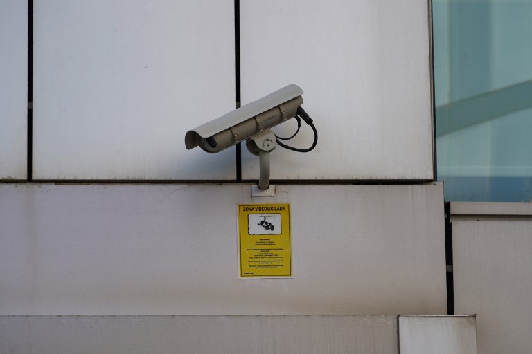 A security camera mounted on a wall above a yellow sign that reads “ZONA VIDEOVIGILADA,” indicating a surveillance area.