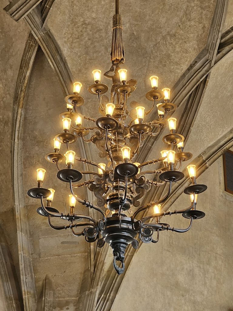 A large, ornate chandelier hanging in St. Vitus Cathedral in Prague castle. The chandelier is made of dark metal and features multiple tiers of lights, with small, warm-toned bulbs illuminating the space.