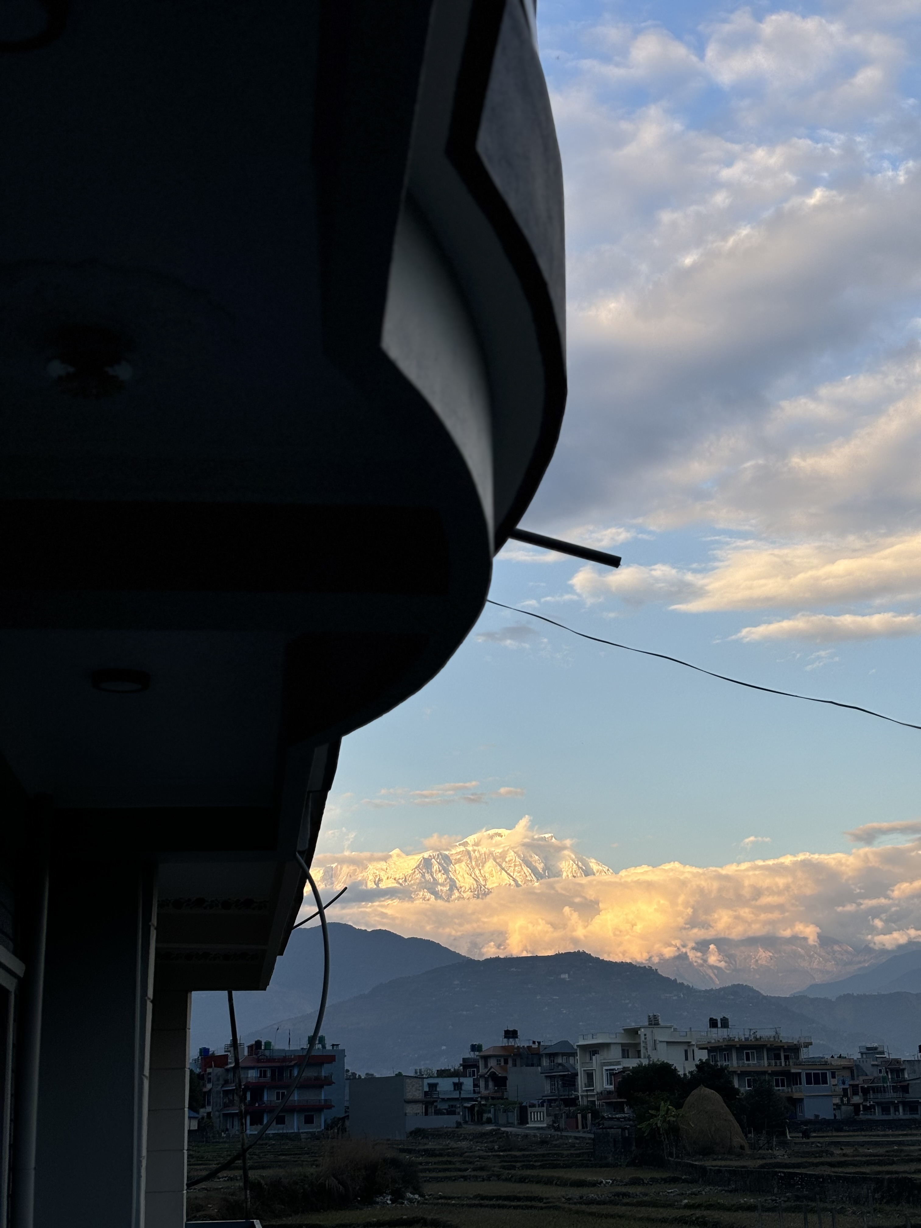 A view of snow-capped mountains in the distance with a partially cloudy sky. In the foreground, a silhouette of a building's balcony and the rooftops of other buildings are visible. The sun casts a warm glow on the mountains and clouds, creating a contrast with the darker foreground.