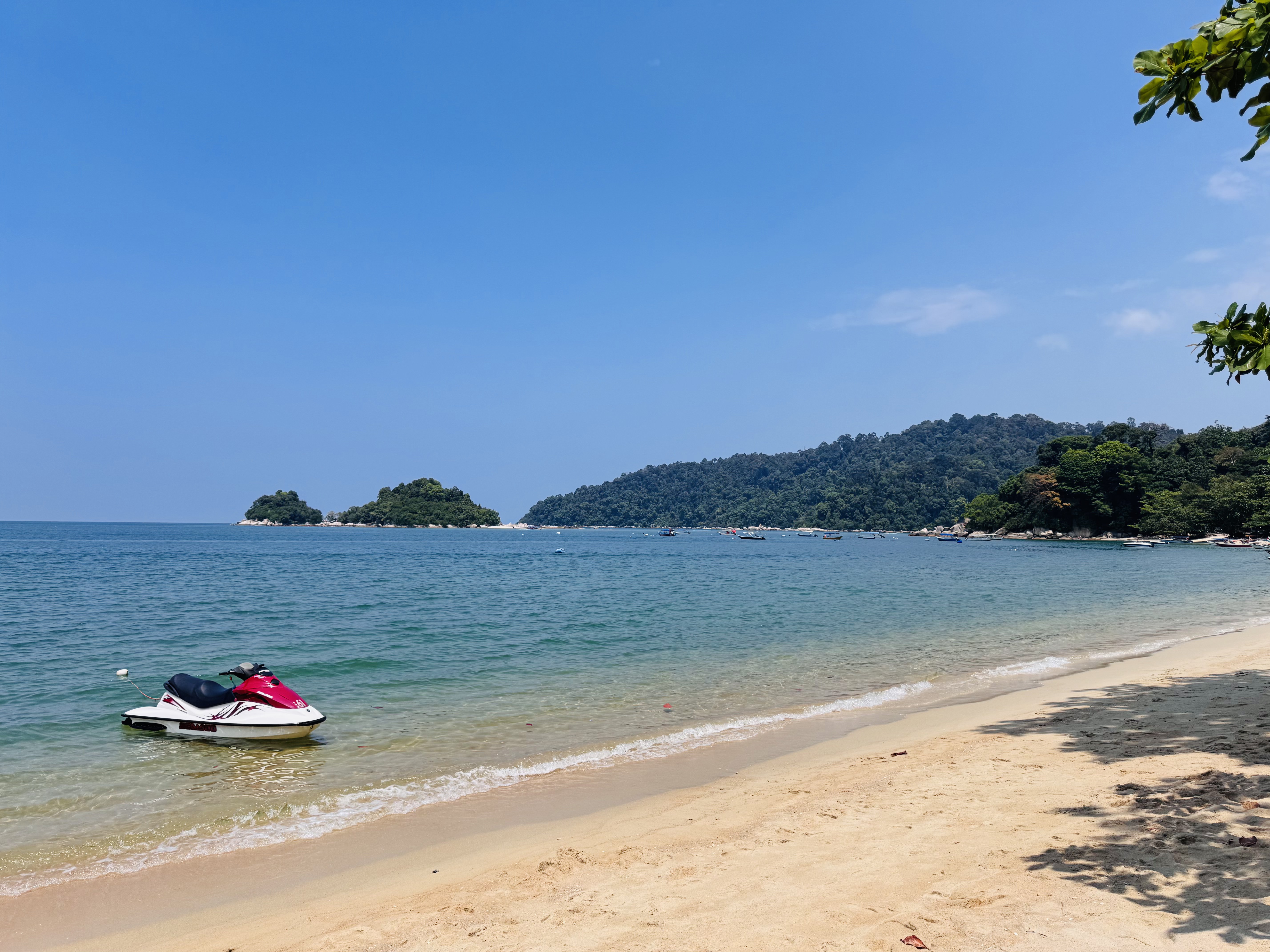 A serene beach scene with clear blue skies and turquoise water. A red and white jet ski is floating near the sandy shore. In the background, there are lush green islands and a forested hillside. Some boats are anchored in the distance.
