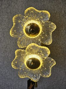 Two flower-shaped wall lamps, one above the other, illuminated against a textured gray background. The lamps have clear, textured glass petals and a warm, glowing center, resembling stylized blossoms. Captured from an electrical shop in Kozhikode.
