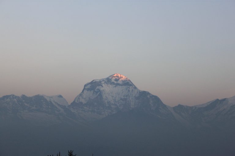 A mountain landscape at dawn with snow-capped peaks. The top of the tallest peak is illuminated by the first light of the sun, giving it a pinkish glow. The sky is clear with subtle hues of blue and gray.