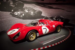 Red vintage race car with the number 7 displayed on the hood and doors, shown on a museum platform with a black-and-white racing photo mural in the background.
