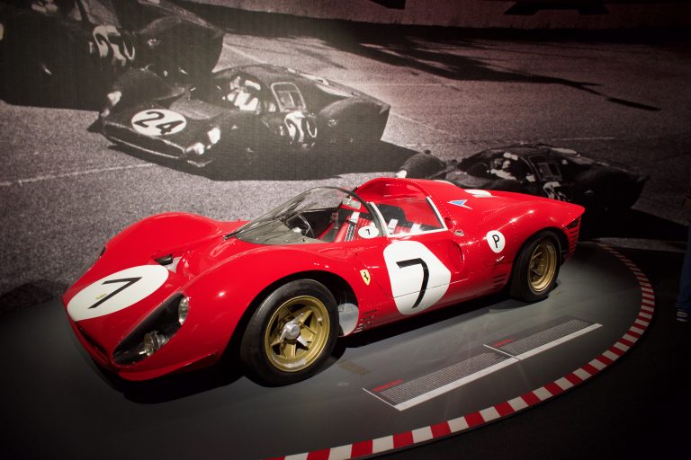 Red vintage race car with the number 7 displayed on the hood and doors, shown on a museum platform with a black-and-white racing photo mural in the background.