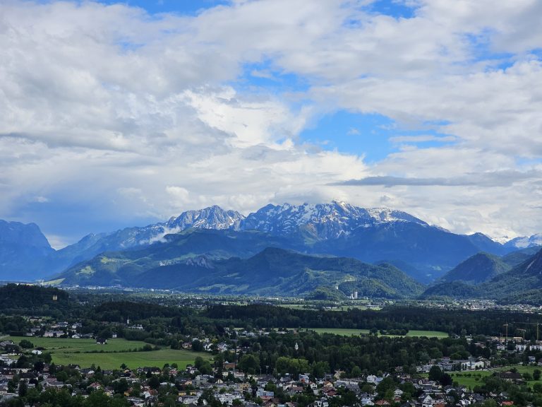 A landscape view of the snow-capped Alps in the distance, with a green forested area and a city in the foreground. The blue sky has scattered white clouds. This stunning view is from Hohensalzburg Fortress in Salzburg, Austria.