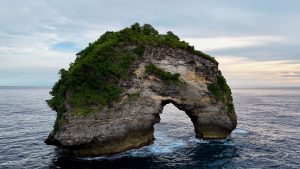 Nusa Banah, a rugged limestone island off the coast of Nusa Penida, featuring a natural rock arch above the ocean, covered in lush green vegetation.
