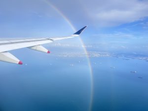 View from an airplane window showing an ocean with numerous ships below and a cityscape in the distance. A partial rainbow arcs across the sky.
