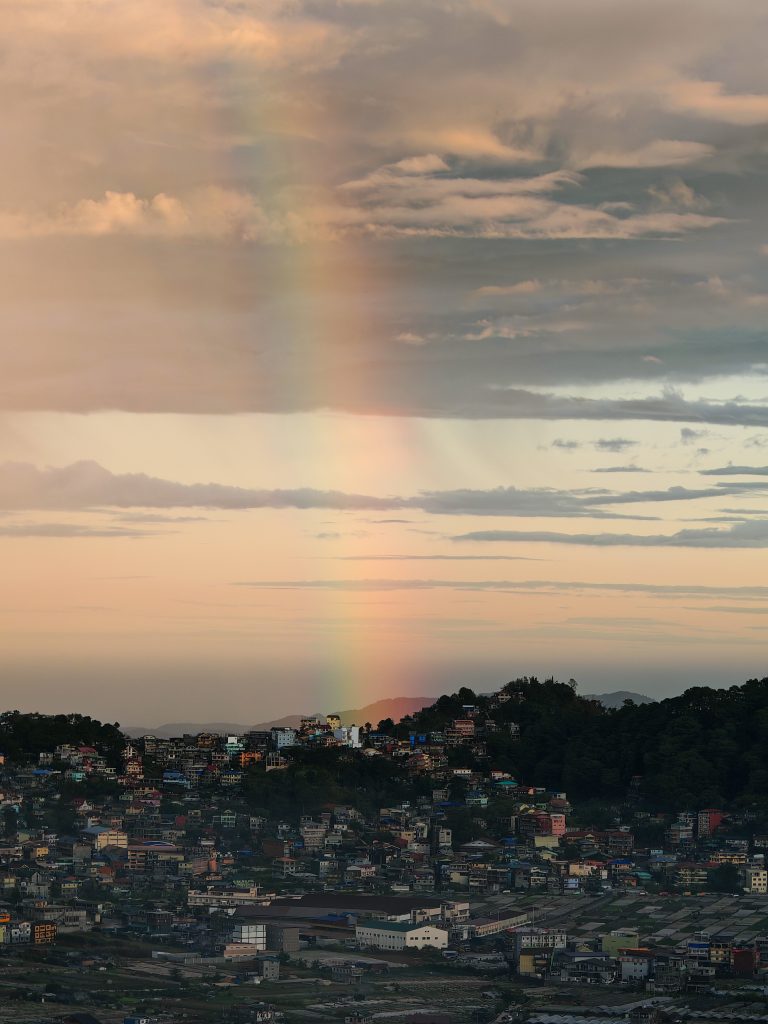 A photo of a sunrise over a cityscape, taken in La Trinidad Benguet. The sky is a mix of pale pink and blue with clouds, hinting at dawn. Below, a densely packed town is visible, silhouetted against the brightening sky. A faint rainbow arc is visible in the distance.