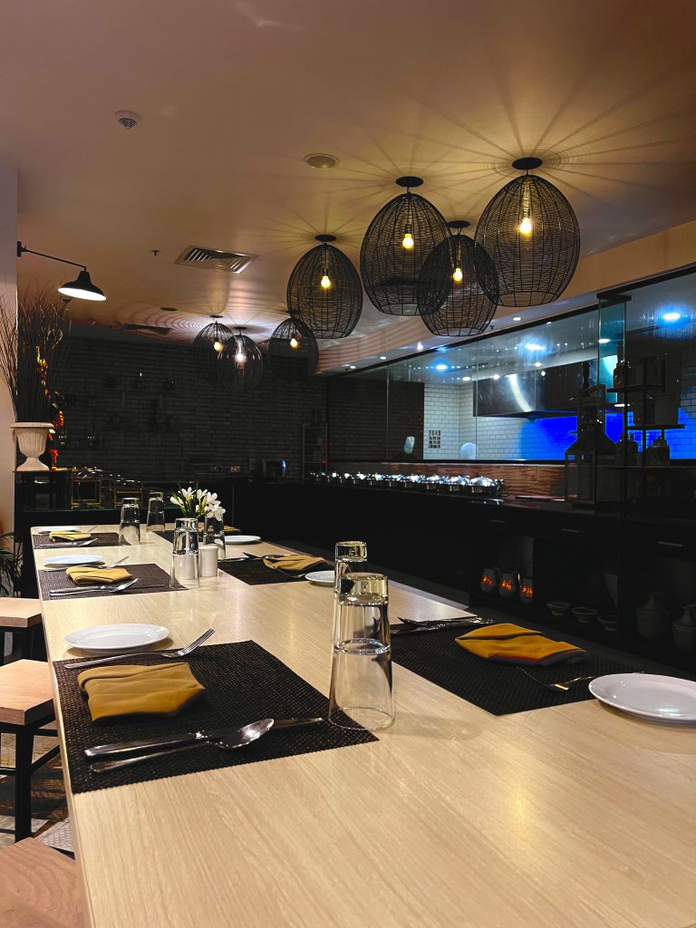 A modern restaurant dining area with a long, light wooden table set for a meal.