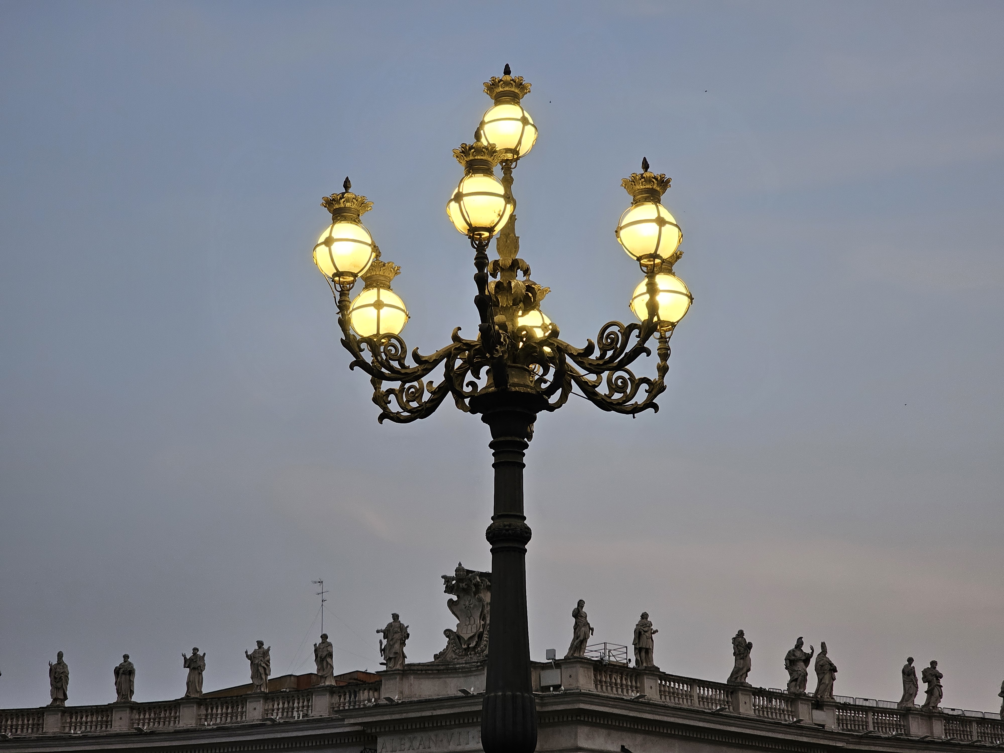 A decorative street lamp with globe-shaped lights, set against a twilight sky at St. Peter's Square in Vatican City. The lamp post is black and ornate, with golden embellishments at the top and around the light fixtures.