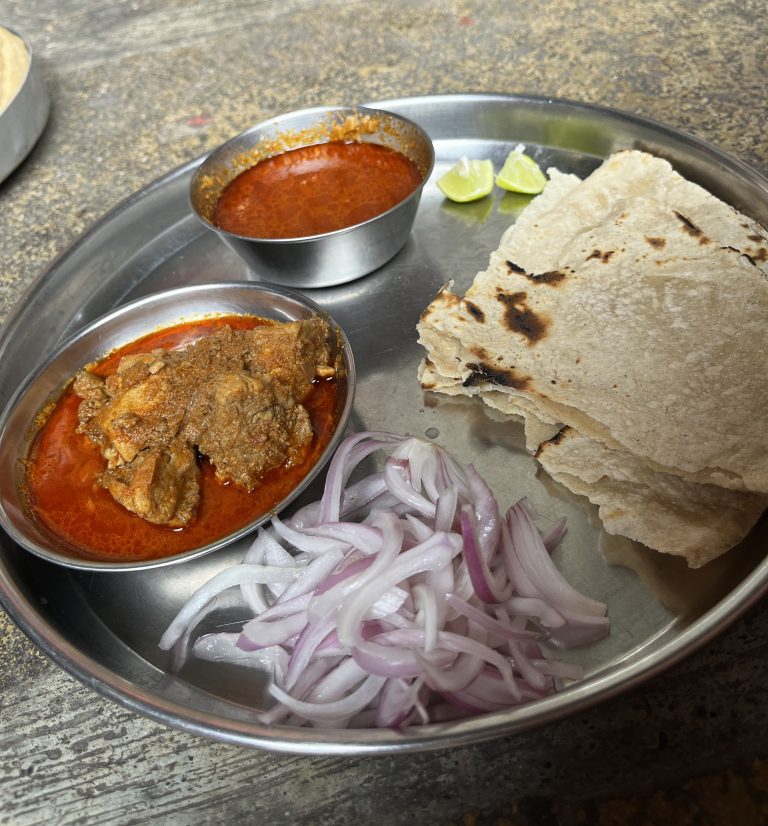 A stainless steel plate with Chulivarchi bhakri, Indian food, consisting on a serving of spicy curry with meat, sliced onions, pieces of flatbread, lime wedges, and a bowl of red curry sauce.