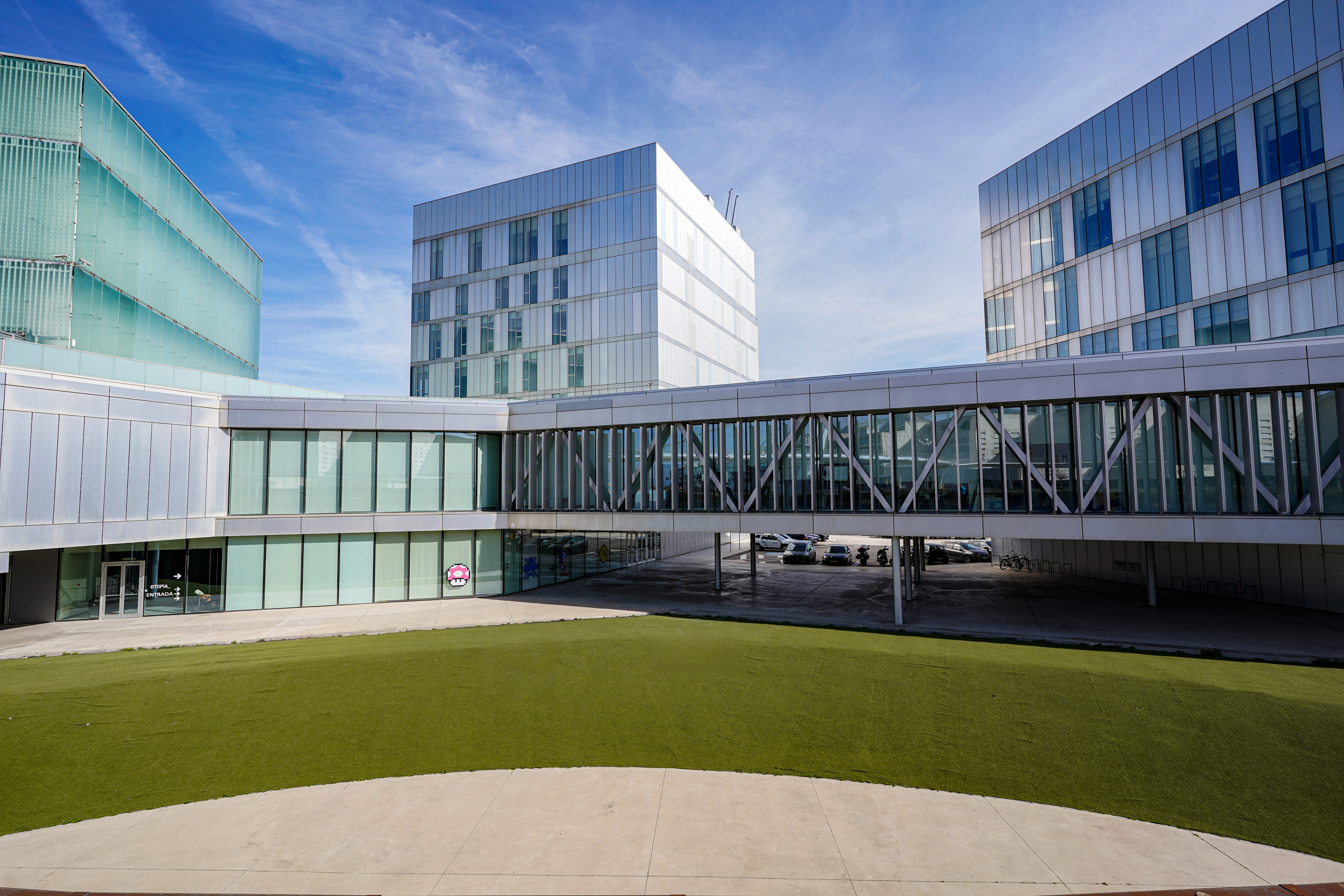 A modern architectural complex featuring three multi-story buildings with glass facades, connected by a sky bridge. The foreground is a courtyard with a circular patch of grass. The sky is clear and blue.