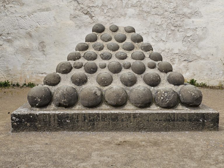 A pyramid-shaped pile of stone balls, likely cannonballs, resting on a stone base. The balls are arranged in rows, decreasing in number towards the top, forming the pyramid shape. The structure is set against a plain, light-colored wall. Located at the Hohensalzburg Fortress in Salzburg, Austria.