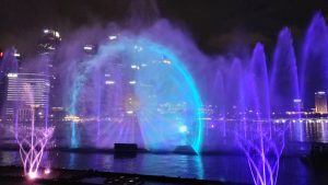 A stunning night view of the Marina Bay Sands Light and Water Show in Singapore, featuring vibrant water fountains illuminated in shades of purple and blue. The mesmerizing water projections create a holographic wheel effect, with the city skyline glowing brightly in the background, adding to the magical ambiance.