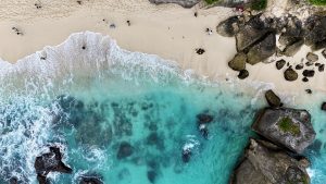 Aerial view of Diamond Beach, Nusa Penida, showing turquoise waves washing onto a sandy shore. Large rocks and cliffs surround the beach, with visitors walking along the shoreline.