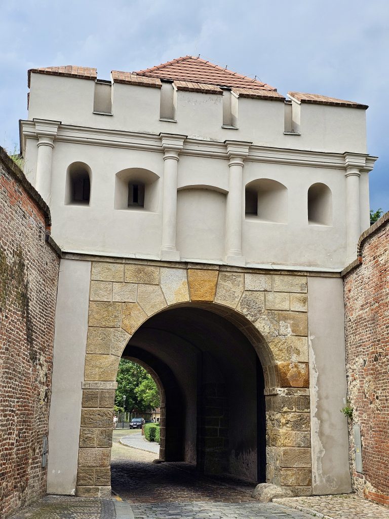 A close view of Táborská brána (Tábor Gate) at Vy?ehrad, Prague. The gate features a stone archway at its base, leading through to the other side, and is topped by a white structure with a red-tiled roof. The gate is flanked by brick walls on either side, and the area around it is paved with cobblestones.