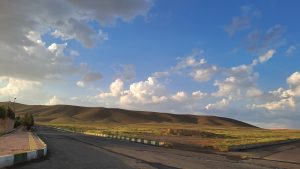 A scenic landscape featuring rolling hills under a blue sky with scattered clouds. In the foreground, there's an asphalt road with a sidewalk lined by trees on the left. The light casts soft shadows on the hills, adding depth to the scene.