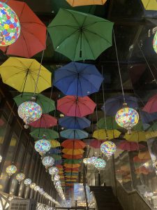 A ceiling decorated with hanging colorful umbrellas and ornate mosaic lamps. The lamps are spherical with intricate patterns, casting a warm glow. The umbrellas are in various colors, including red, green, blue, and yellow.
