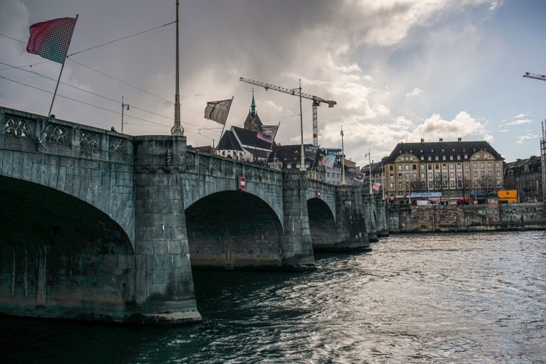 A stone bridge spans across Rhine river with colorful flags attached to it. In the background, construction cranes and a historic building can be seen under a partly cloudy sky.