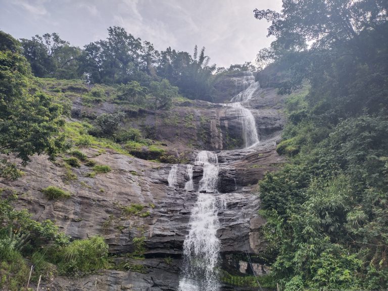 A cascading waterfall flows down a rocky cliff surrounded by lush green vegetation and trees under a cloudy sky.