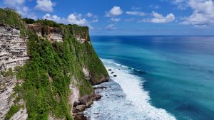 A coastal view of the towering limestone cliffs near Uluwatu Temple in Bali. The deep blue ocean stretches to the horizon, with waves crashing against the rocky shoreline below.