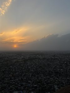 Aerial view of a cityscape at sunset, with the sun partially obscured by clouds. The sky displays a gradient of warm hues, while the densely packed buildings below gradually fade into the horizon.