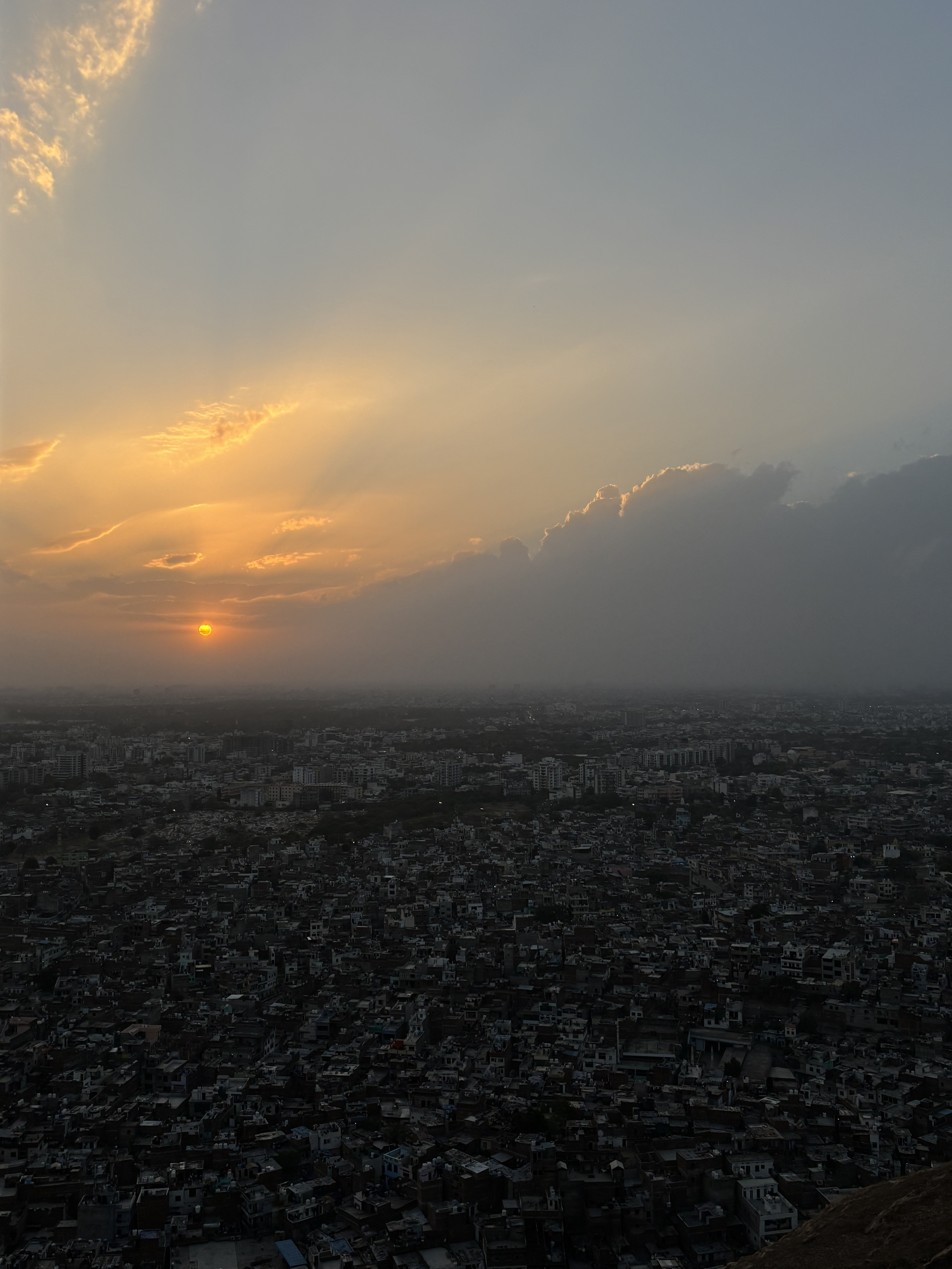 Aerial view of a cityscape at sunset, with the sun partially obscured by clouds. The sky displays a gradient of warm hues, while the densely packed buildings below gradually fade into the horizon.