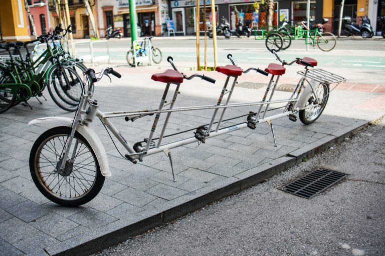 A long tandem bike with four seats and handlebars, each seat has a red saddle. The bike is parked on a paved sidewalk with other bicycles and motorcycles in the background.