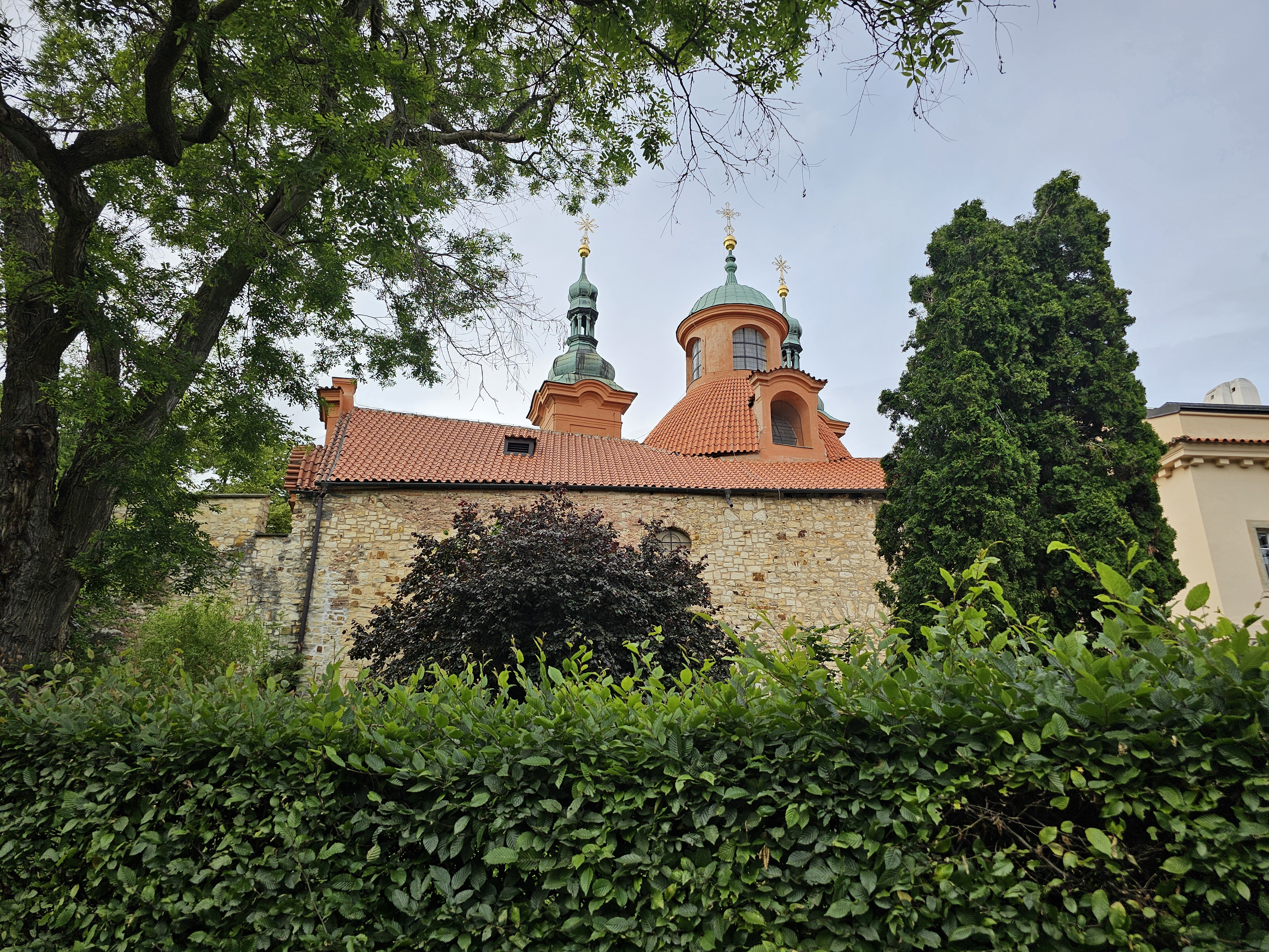 A long view of the Cathedral of St. Lawrence in Prague, with its red-tiled roofs and green-domed towers, partially obscured by lush greenery. A stone wall forms the base of the cathedral, while trees and a neatly trimmed hedge fill the foreground. The sky is overcast, providing a soft, diffused light to the scene.